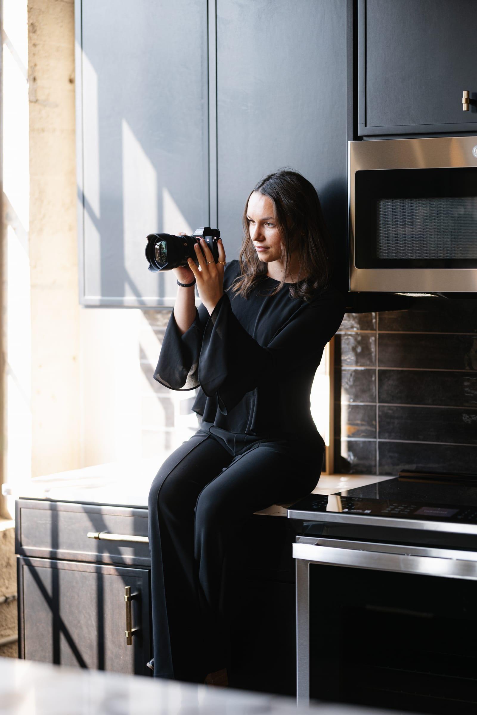 Ashlin seated on a kitchen counter, camera raised, shooting toward hard window light