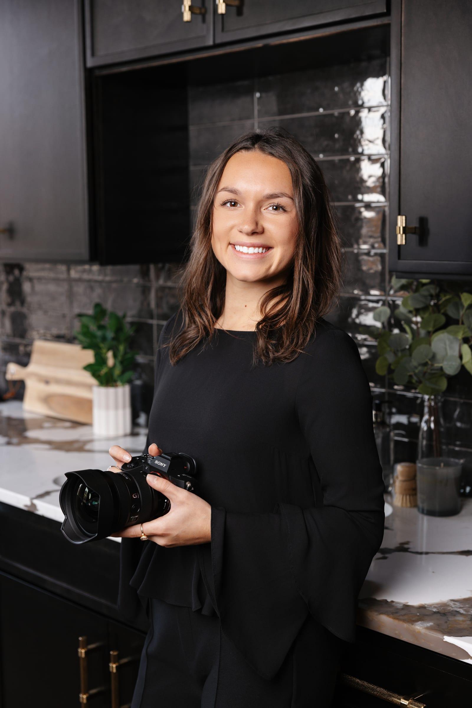 Ashlin Samples smiling, holding a Sony camera in a dark designer kitchen