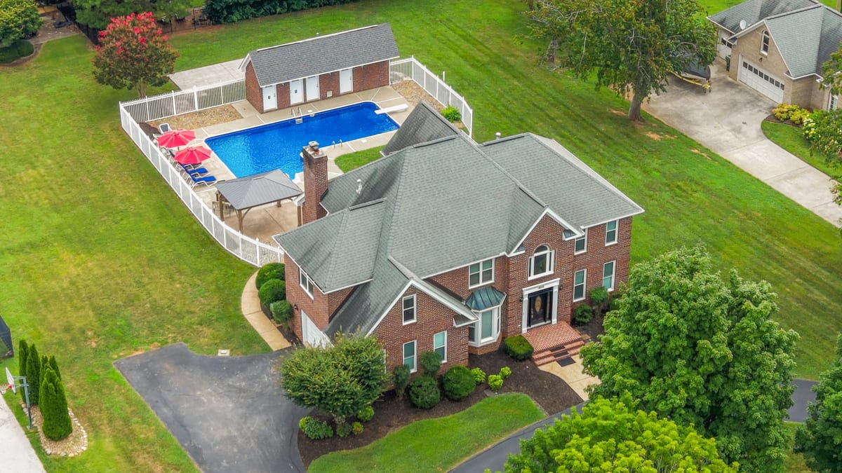 Drone aerial of a brick estate with fenced pool, pool house, and broad lawns