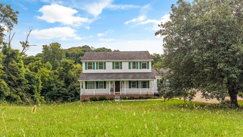 White colonial with green shutters and wraparound porch on an open hillside lawn